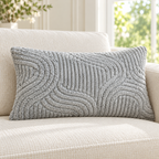 A rectangular gray textured lumbar pillow with a raised wavy pattern, resting on a cream-colored sofa in a bright, modern living room.