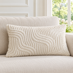 A rectangular cream throw pillow with a raised wavy textured pattern, resting on a matching beige fabric sofa in a bright, modern living room setting.