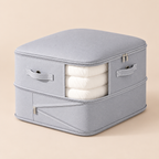A light gray fabric storage bag with a zippered top and side pockets, shown holding three folded white sheets or blankets. The bag has fabric handles and silver zipper pulls, photographed on a plain beige studio background.