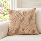 A square beige throw pillow with a textured wavy cable knit pattern, resting on a cream-colored sofa in front of a window.