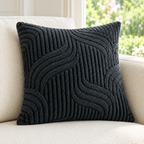 A square dark charcoal gray textured throw pillow with a raised wavy pattern, resting on a light beige upholstered sofa with a softly blurred window background.