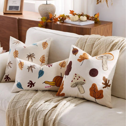 Three decorative throw pillows with an autumn-themed embroidered pattern of leaves, acorns, and mushrooms on a cream background, styled on a white sofa with a beige knit throw blanket.