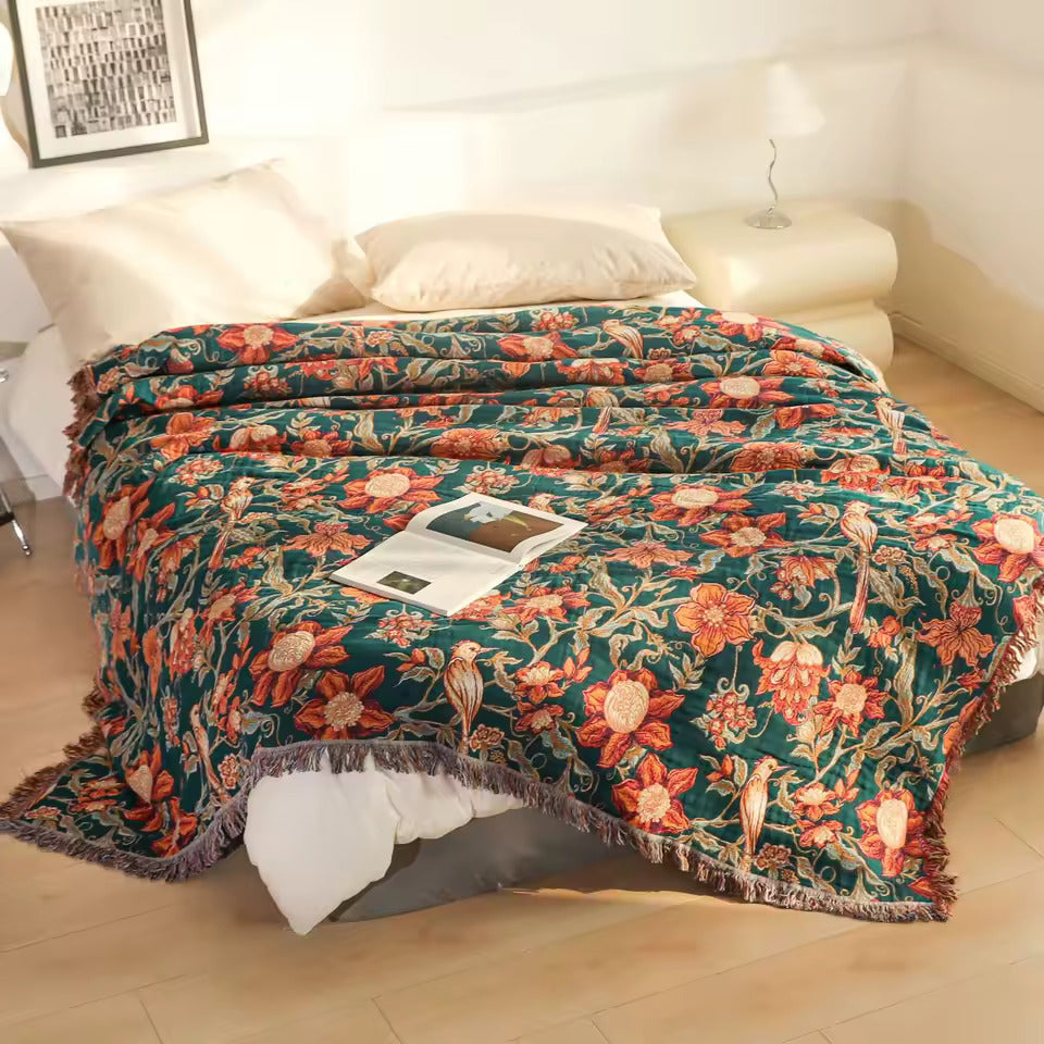 A floral throw blanket draped over a bed in a softly lit bedroom. The blanket has a dark teal-green background with large orange, red, and cream flowers, green leaves, and small bird motifs, plus fringed edges.