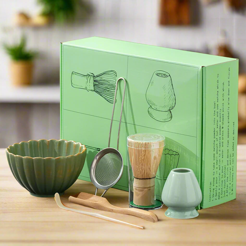 A set of traditional Japanese matcha tea preparation tools displayed in front of their packaging box, including a blue matcha bowl, a bamboo whisk, a bamboo rest, a metal sifting bowl, and two small blue cups.
