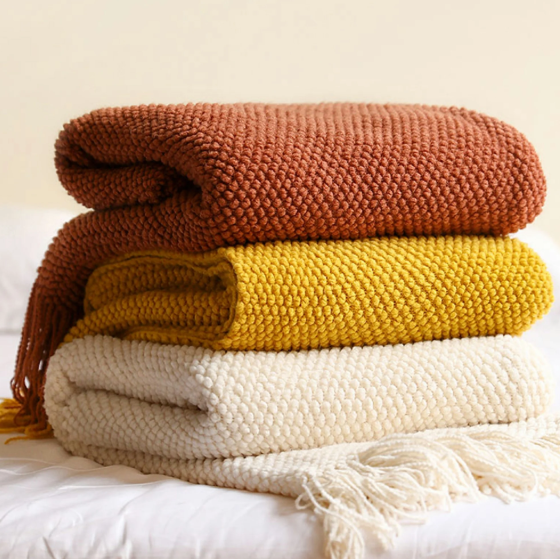 Three folded chunky knit throw blankets stacked on a white bed: a rust orange top blanket, a mustard yellow middle blanket, and an off-white cream bottom blanket with fringe.