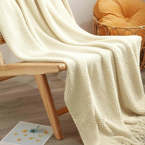 A cream-colored waffle-knit throw blanket with fringe, draped over a light wood chair in a cozy room setting with a yellow cushion in the background.