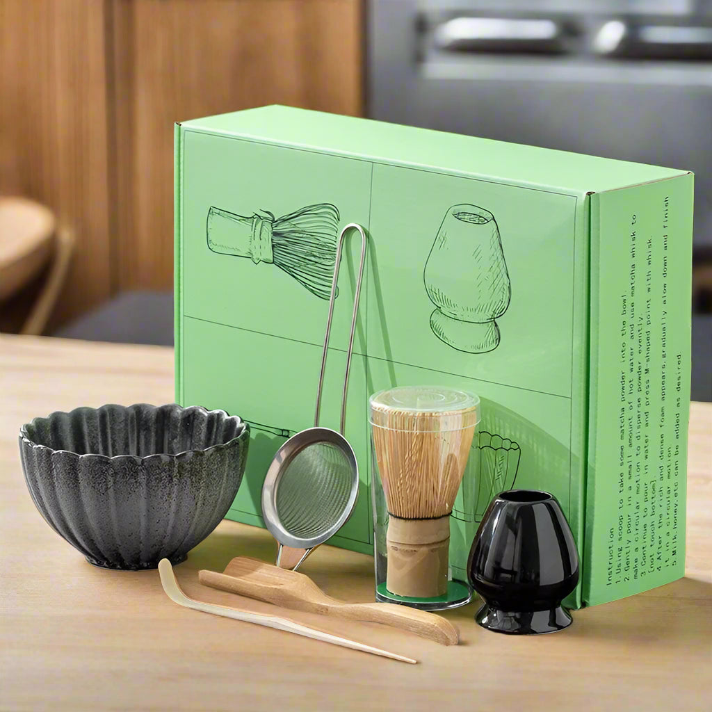 A set of traditional Japanese matcha tea preparation tools displayed in front of their packaging box, including a blue matcha bowl, a bamboo whisk, a bamboo rest, a metal sifting bowl, and two small blue cups.