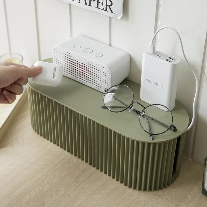 A beige cable and charger organizer on a desk with a smartphone on top and a white mouse connected to it, placed next to a lamp and a bottle.