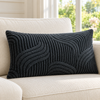 A rectangular lumbar throw pillow with a dark charcoal gray textured wavy pattern, resting on a cream-colored sofa in a bright, modern living room.