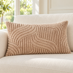 A rectangular beige throw pillow with a textured, raised wavy pattern, resting on a cream-colored sofa in a bright, sunlit room.