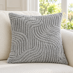 A square gray textured throw pillow with a raised wavy cable knit pattern, resting on a light beige upholstered armchair in a bright living room setting.